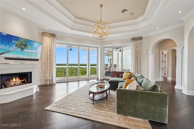 living room featuring an inviting chandelier, a tray ceiling, crown molding, dark wood-type flooring, and ornate columns