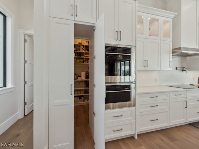 kitchen with white cabinets, wall chimney exhaust hood, black electric cooktop, double oven, and light wood-type flooring