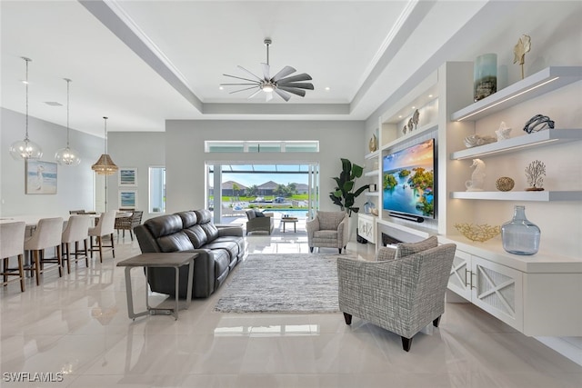 living room with ceiling fan, ornamental molding, a fireplace, a tray ceiling, and built in features