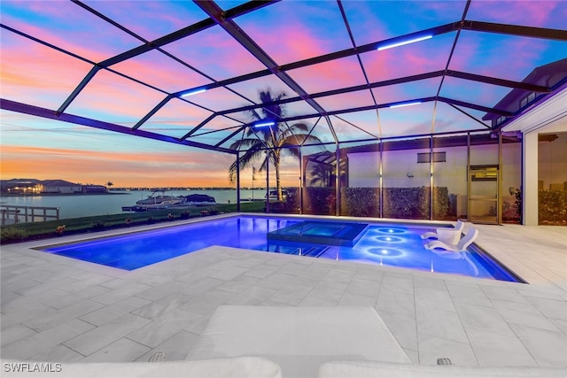 pool at dusk featuring a patio area, glass enclosure, an in ground hot tub, and a water view