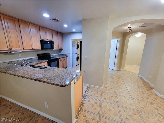 kitchen with dark stone counters, black appliances, kitchen peninsula, and stacked washing maching and dryer