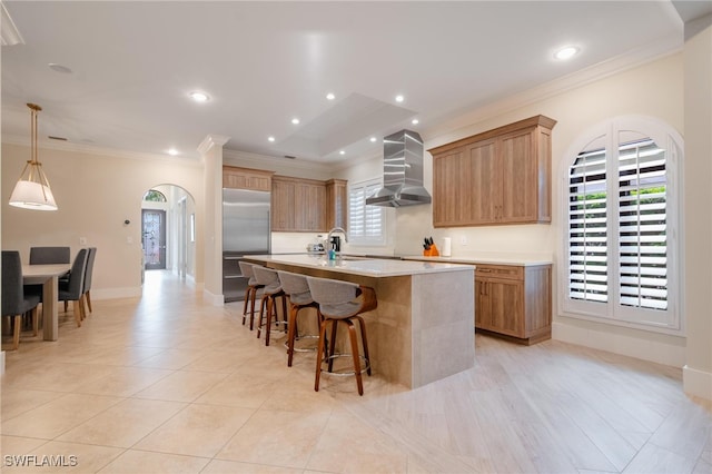 kitchen with sink, an island with sink, stainless steel built in refrigerator, wall chimney exhaust hood, and decorative light fixtures