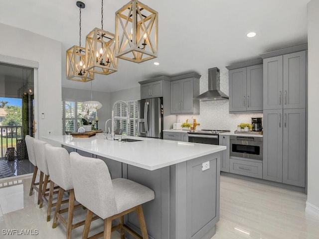 kitchen with a sink, an inviting chandelier, stainless steel appliances, gray cabinets, and wall chimney exhaust hood