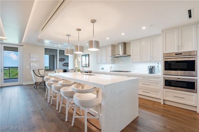 kitchen with white cabinets, wall chimney range hood, an island with sink, decorative light fixtures, and dark hardwood / wood-style flooring