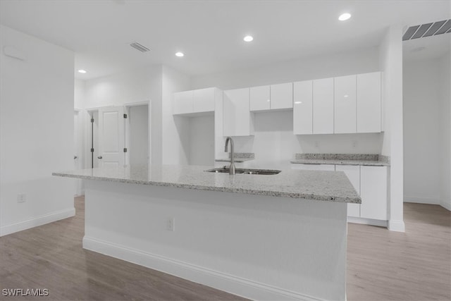 kitchen with light wood-type flooring, a kitchen island with sink, sink, white cabinets, and light stone countertops