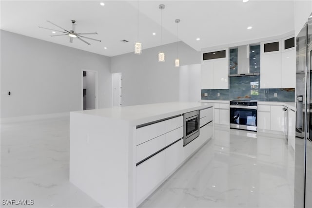 kitchen featuring white cabinets, stainless steel appliances, a center island, ceiling fan, and wall chimney range hood