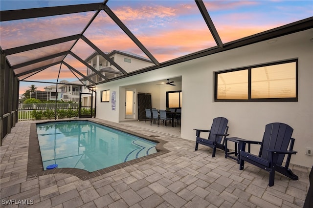 pool at dusk featuring glass enclosure, ceiling fan, and a patio area