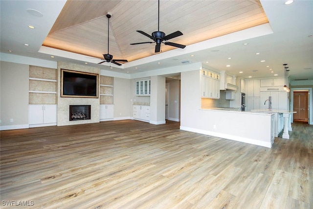 unfurnished living room featuring light wood-type flooring, a tray ceiling, a large fireplace, and ceiling fan