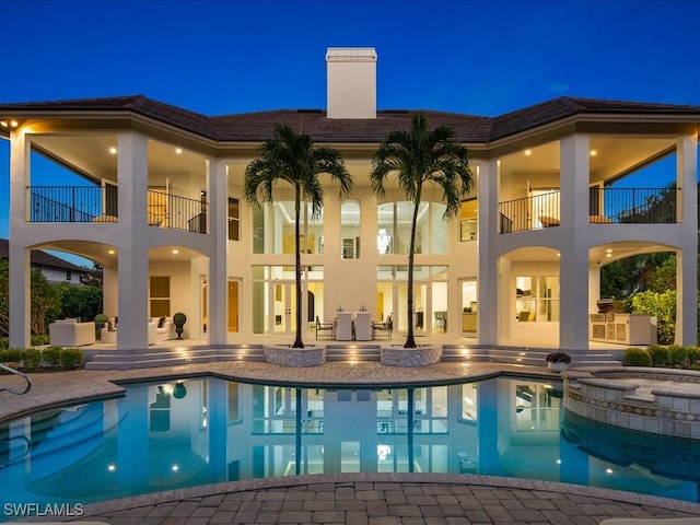 back house at twilight with a balcony, a swimming pool with hot tub, and a patio area