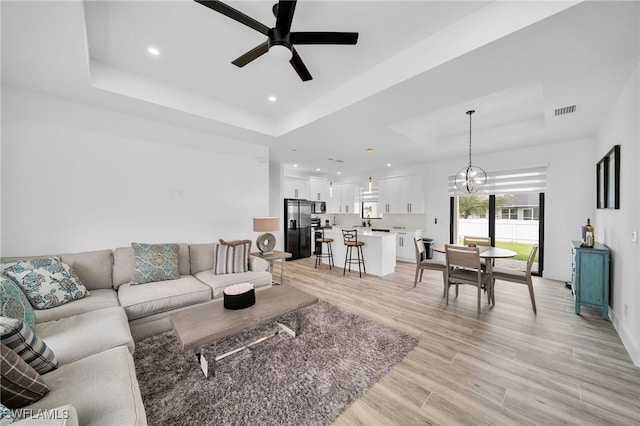 living room featuring ceiling fan with notable chandelier, light wood-type flooring, and a raised ceiling