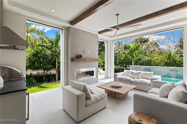 living area with a healthy amount of sunlight, ceiling fan, light tile patterned floors, beamed ceiling, and a glass covered fireplace