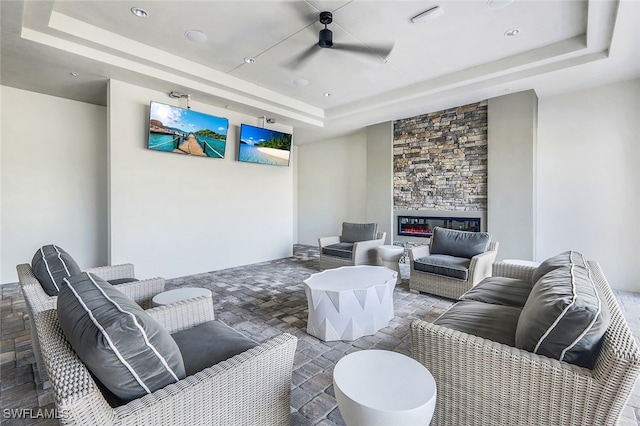 living room with a raised ceiling, a stone fireplace, and ceiling fan