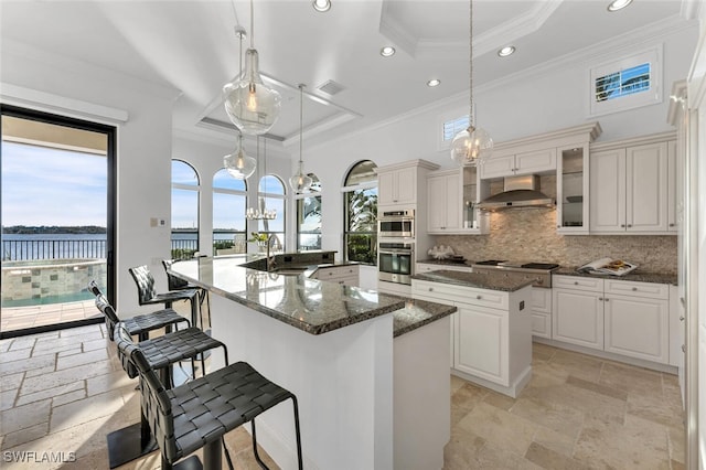 kitchen featuring a spacious island, wall chimney range hood, decorative light fixtures, a water view, and a raised ceiling