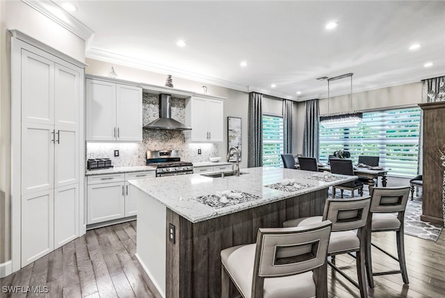 kitchen featuring wall chimney exhaust hood, sink, white cabinetry, stainless steel range with gas cooktop, and an island with sink