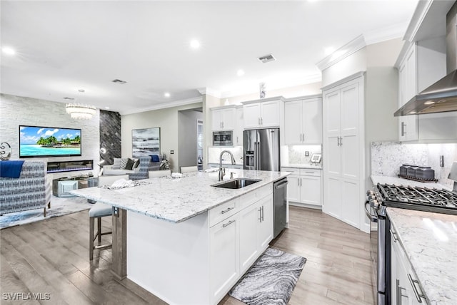 kitchen featuring appliances with stainless steel finishes, sink, white cabinets, wall chimney range hood, and a center island with sink