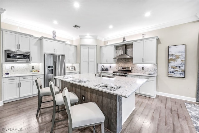 kitchen with appliances with stainless steel finishes, sink, a kitchen island with sink, light stone counters, and wall chimney exhaust hood