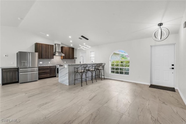 kitchen featuring lofted ceiling, a kitchen island with sink, a kitchen breakfast bar, wall chimney exhaust hood, and appliances with stainless steel finishes