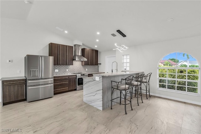 kitchen with a kitchen bar, wall chimney exhaust hood, dark brown cabinets, stainless steel appliances, and hanging light fixtures