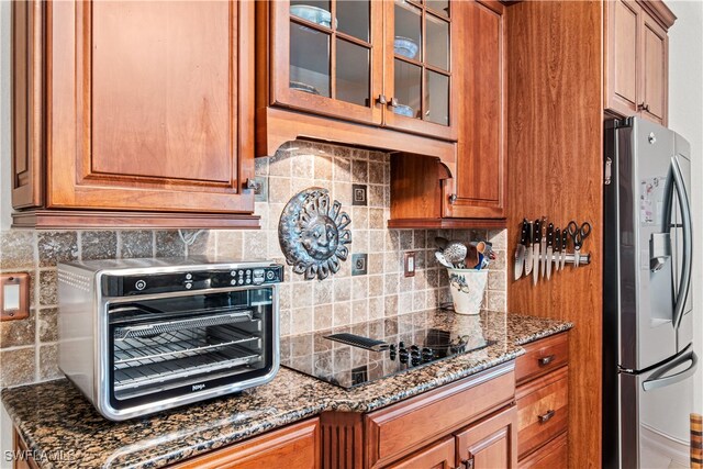 kitchen featuring black electric cooktop, glass insert cabinets, stainless steel refrigerator with ice dispenser, and dark stone counters