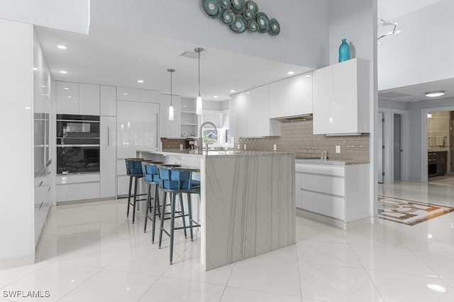 kitchen with a center island with sink, decorative light fixtures, backsplash, white cabinetry, and double oven