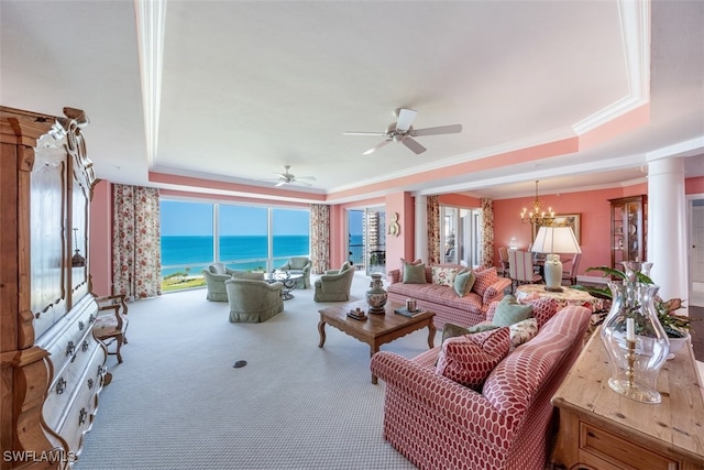 carpeted living room featuring decorative columns, ornamental molding, ceiling fan with notable chandelier, a tray ceiling, and a water view