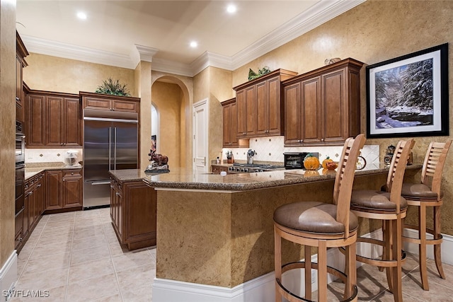 kitchen featuring stainless steel built in fridge, dark stone counters, a center island, ornamental molding, and a breakfast bar