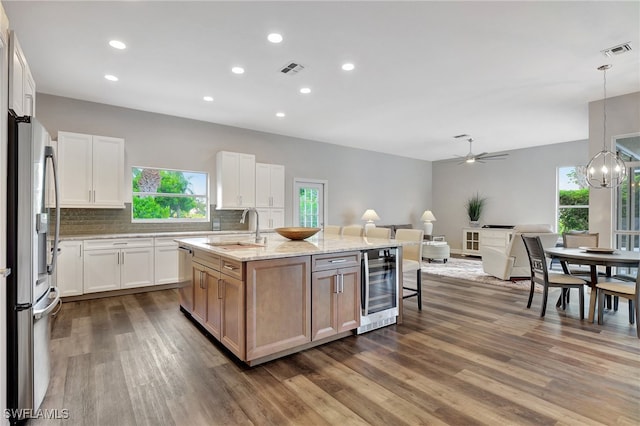 kitchen featuring white cabinets, ceiling fan with notable chandelier, light stone countertops, beverage cooler, and a center island with sink