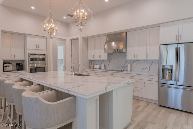 kitchen featuring appliances with stainless steel finishes, a chandelier, a center island with sink, sink, and wall chimney range hood