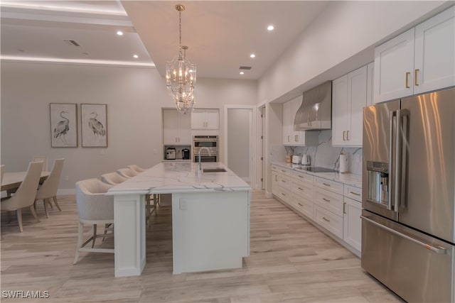 kitchen featuring a kitchen island with sink, wall chimney exhaust hood, a chandelier, white cabinetry, and appliances with stainless steel finishes