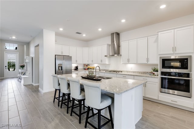 kitchen featuring an island with sink, white cabinetry, appliances with stainless steel finishes, and wall chimney range hood