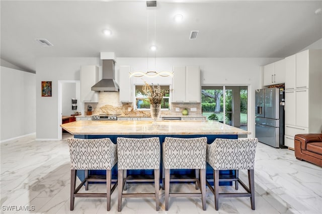 kitchen featuring tasteful backsplash, decorative light fixtures, wall chimney exhaust hood, stainless steel refrigerator with ice dispenser, and a breakfast bar area