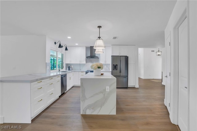 kitchen featuring white cabinetry, stainless steel appliances, wall chimney range hood, decorative light fixtures, and light wood-type flooring