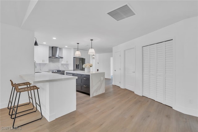 kitchen with kitchen peninsula, wall chimney exhaust hood, stainless steel appliances, decorative light fixtures, and white cabinets