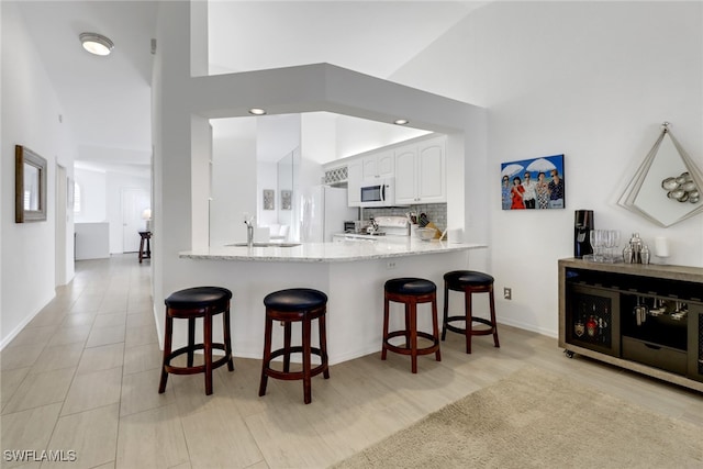 kitchen featuring kitchen peninsula, a kitchen bar, white cabinetry, light stone counters, and white appliances