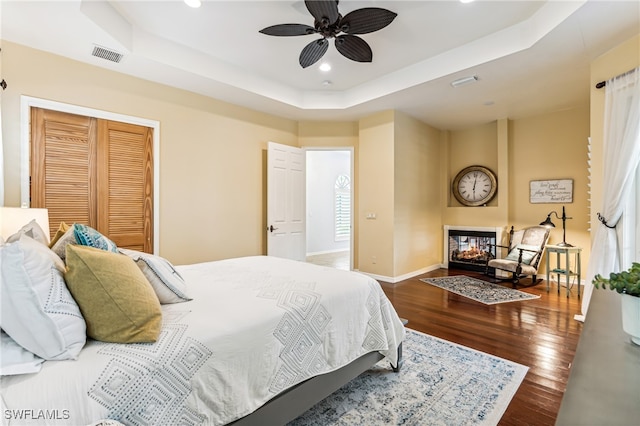 bedroom featuring a multi sided fireplace, a tray ceiling, dark hardwood / wood-style flooring, and a closet