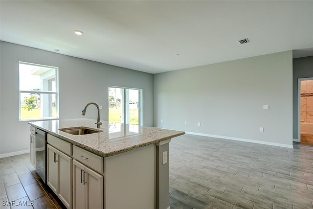 kitchen with sink, stainless steel dishwasher, a kitchen island with sink, light hardwood / wood-style flooring, and light stone countertops