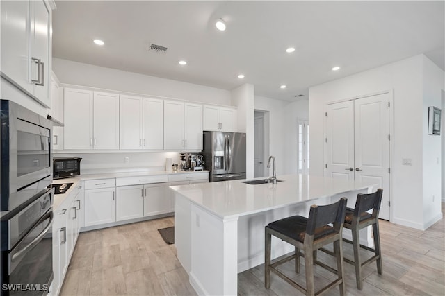 kitchen with white cabinets, appliances with stainless steel finishes, a kitchen island with sink, light hardwood / wood-style floors, and sink