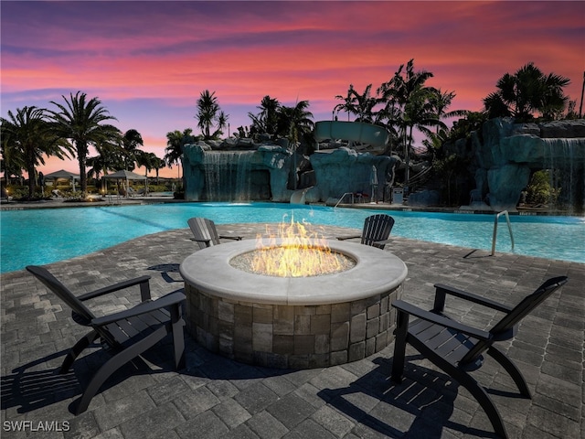 pool at dusk featuring pool water feature, a patio, and an outdoor fire pit