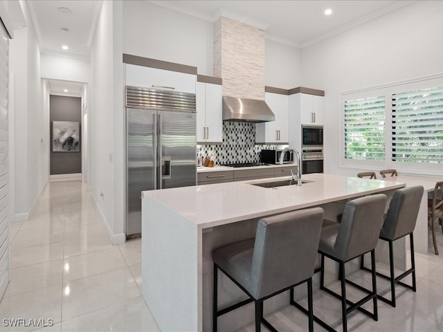 kitchen with white cabinetry, an island with sink, built in appliances, sink, and wall chimney range hood