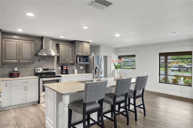 kitchen with wall chimney exhaust hood, appliances with stainless steel finishes, light countertops, an island with sink, and baseboards