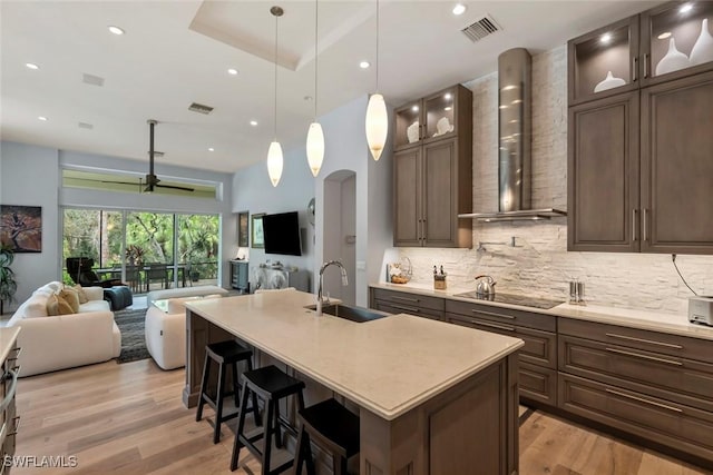 kitchen with a center island with sink, wall chimney range hood, sink, black electric cooktop, and light hardwood / wood-style floors