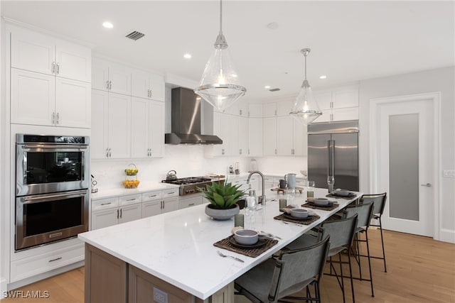 kitchen featuring wall chimney exhaust hood, stainless steel appliances, white cabinetry, and a kitchen island with sink