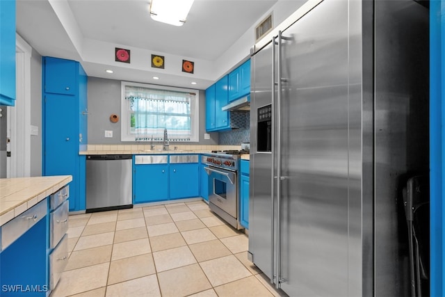 kitchen with blue cabinets, light tile patterned floors, premium appliances, backsplash, and tile countertops