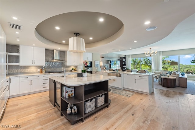 kitchen with light hardwood / wood-style floors, an island with sink, white cabinetry, wall chimney range hood, and a raised ceiling