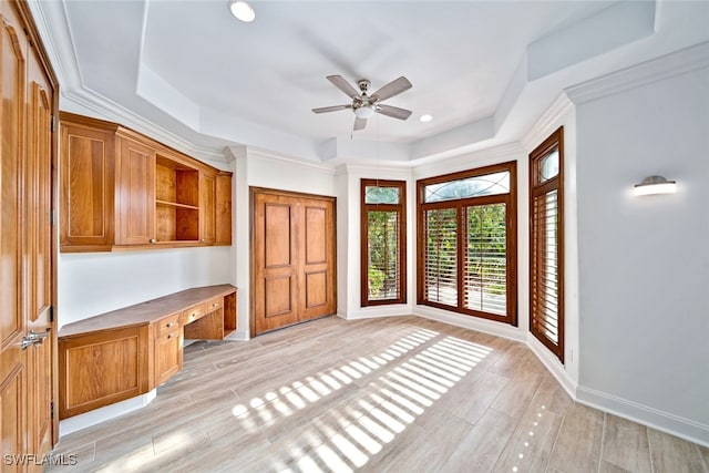 unfurnished office with light wood-type flooring, built in desk, a raised ceiling, ceiling fan, and ornamental molding