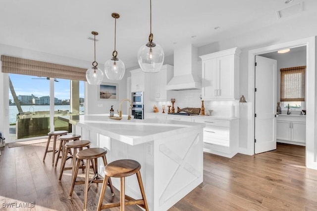 kitchen featuring white cabinetry, premium range hood, hanging light fixtures, a kitchen island with sink, and a water view