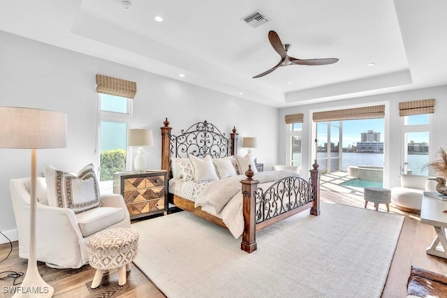 bedroom featuring hardwood / wood-style floors, access to exterior, a water view, ceiling fan, and a tray ceiling