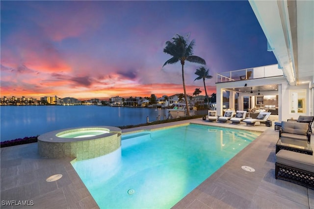 pool at dusk featuring a water view, ceiling fan, a patio area, and an in ground hot tub