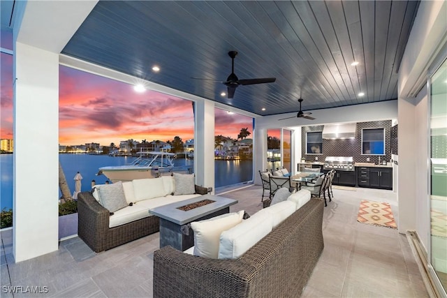 patio terrace at dusk featuring exterior kitchen, an outdoor living space with a fire pit, a water view, ceiling fan, and area for grilling