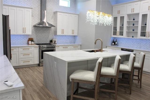 kitchen featuring white cabinets, appliances with stainless steel finishes, a kitchen island with sink, and wall chimney range hood